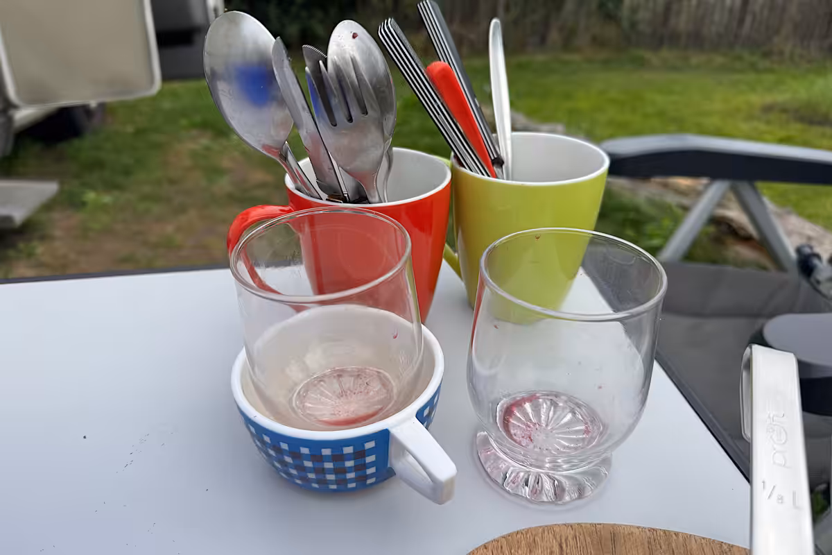Besteck in bunten Bechern und zwei Gläser auf einem Campingtisch