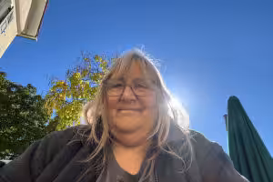 Andrea Sam lächelt im Gegenlicht unter blauem Himmel, daneben ein Baum und ein grüner Sonnenschirm